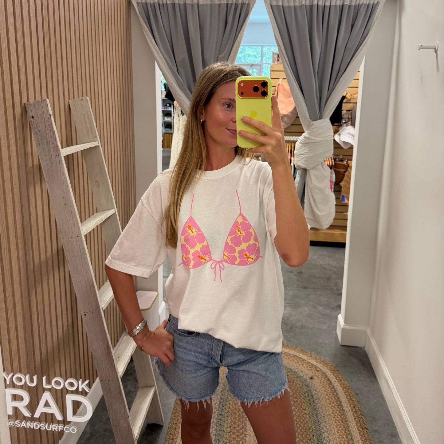 A woman takes a mirror selfie in a boutique wearing a white graphic t-shirt with a pink and yellow bikini top design, paired with denim shorts. The boutique interior features wood-paneled walls, a wooden ladder, gray curtains, and a patterned rug.