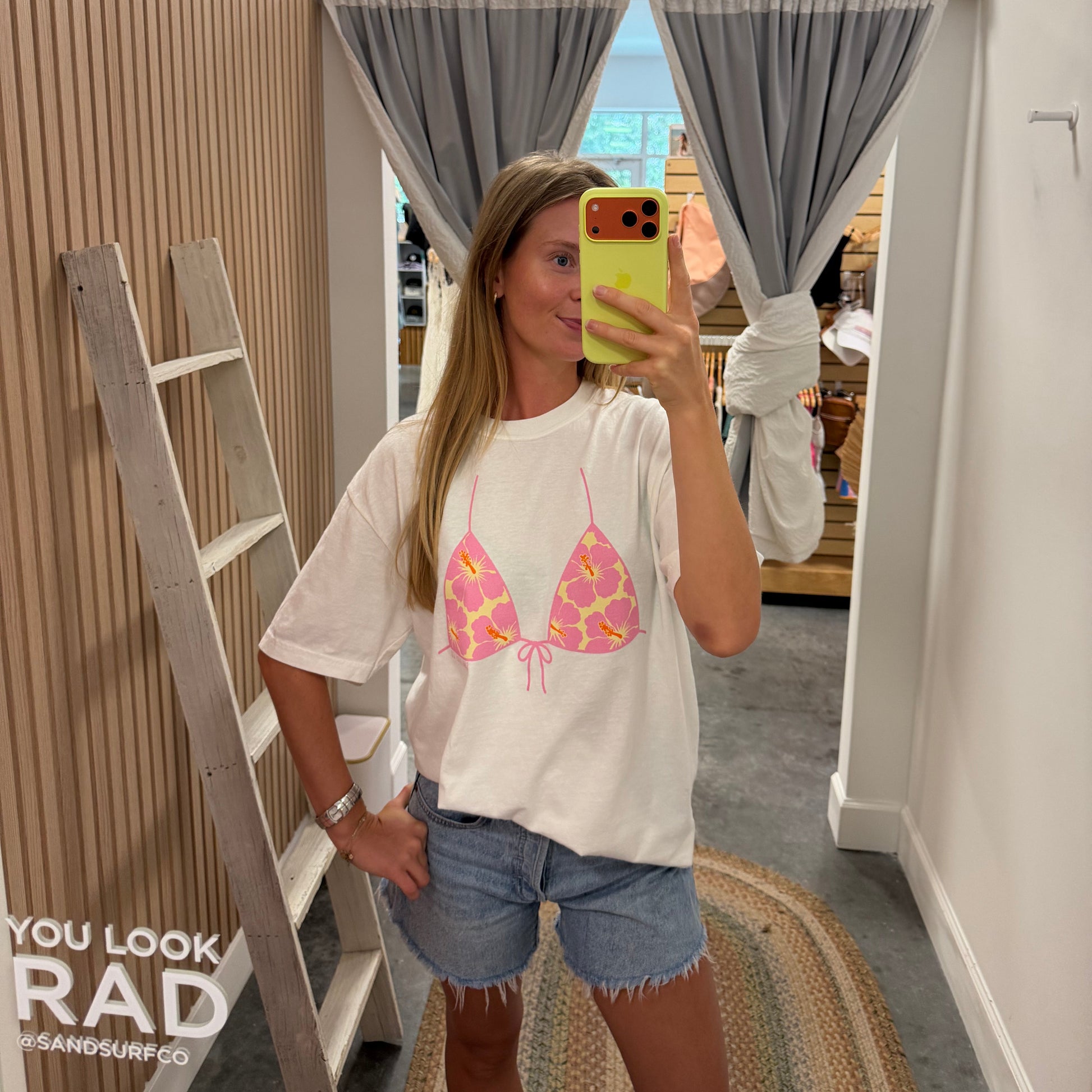 A woman takes a mirror selfie in a boutique wearing a white graphic t-shirt with a pink and yellow bikini top design, paired with denim shorts. The boutique interior features wood-paneled walls, a wooden ladder, gray curtains, and a patterned rug.