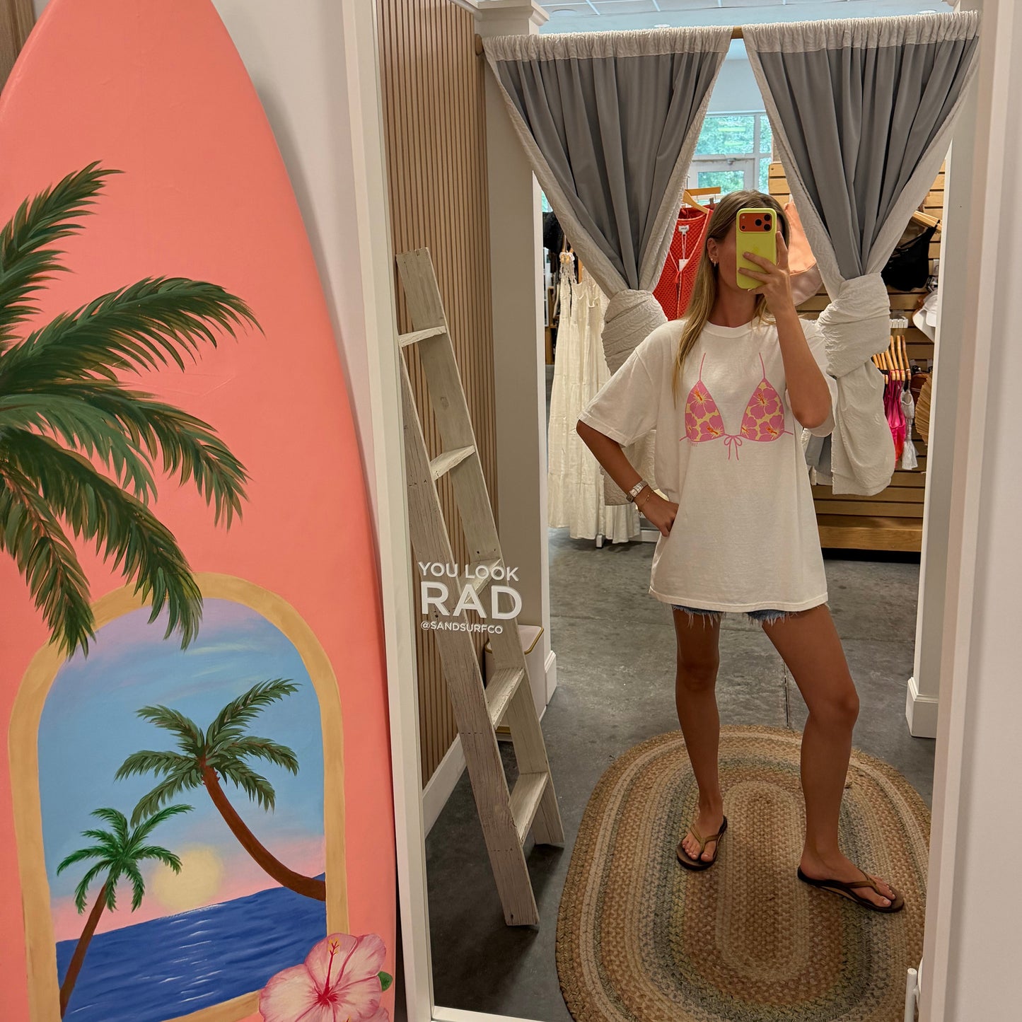 A woman takes a mirror selfie in a boutique showing a white tunic top with a pink bikini-style print. A coral surfboard with a tropical beach illustration leans against the wall on the left.