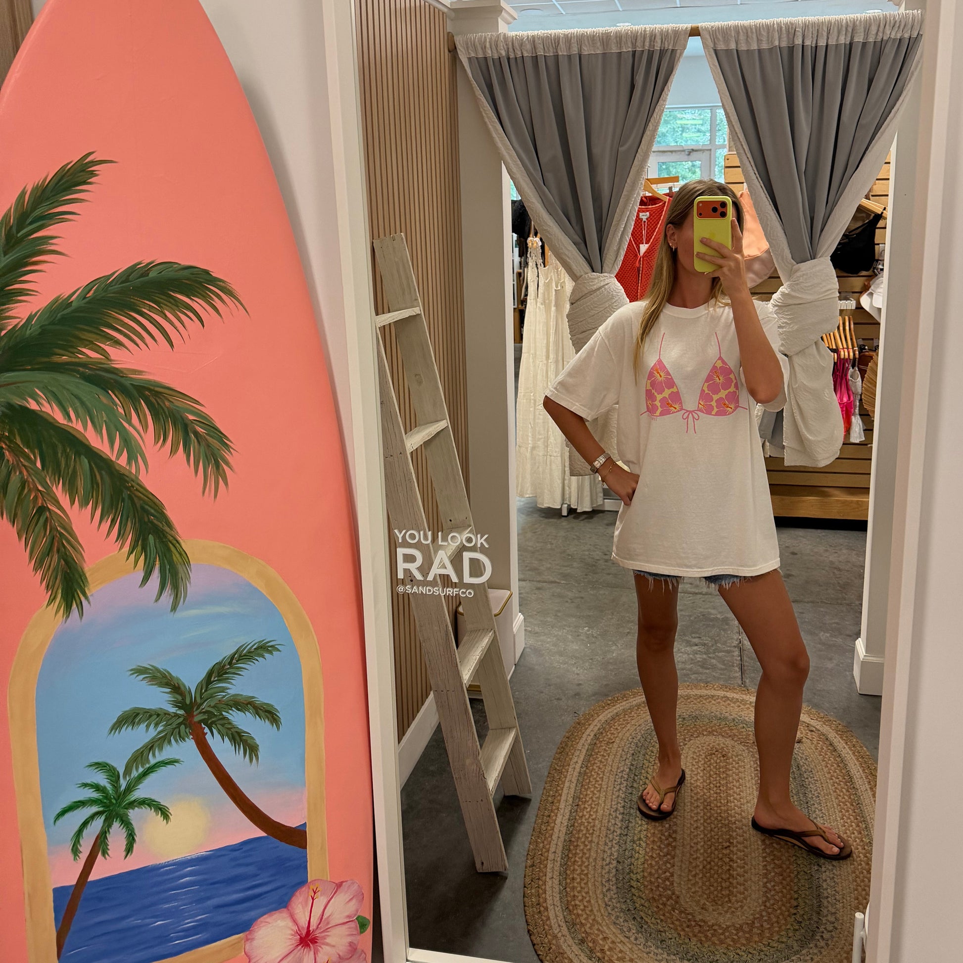 A woman takes a mirror selfie in a boutique showing a white tunic top with a pink bikini-style print. A coral surfboard with a tropical beach illustration leans against the wall on the left.